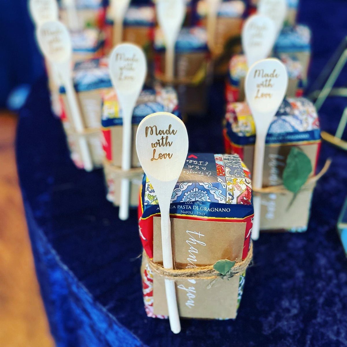 Small pasta gift packages with engraved wooden spoons on a blue velvet cloth.