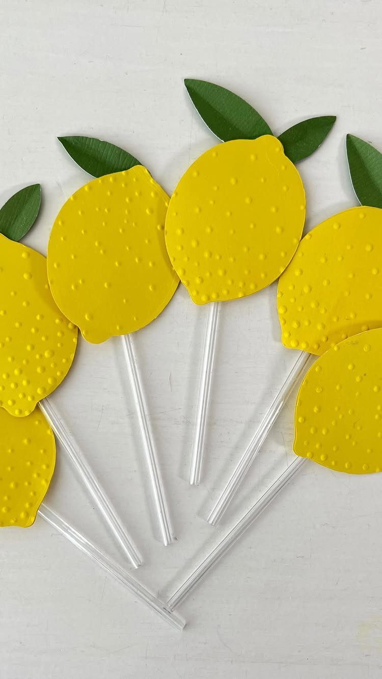 Yellow lemon-shaped cutouts with green leaves on clear plastic sticks on white wood.