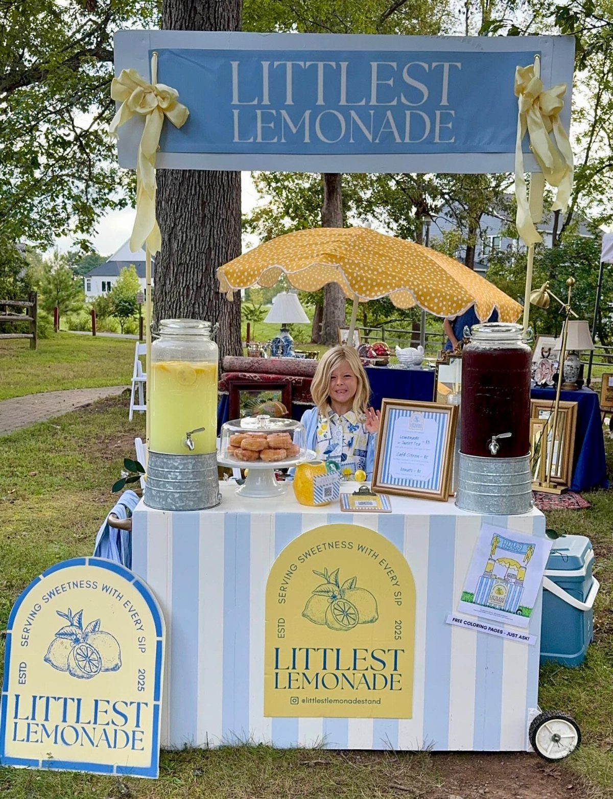 Young girl behind a decorated lemonade stand with drink dispensers and yellow umbrella.