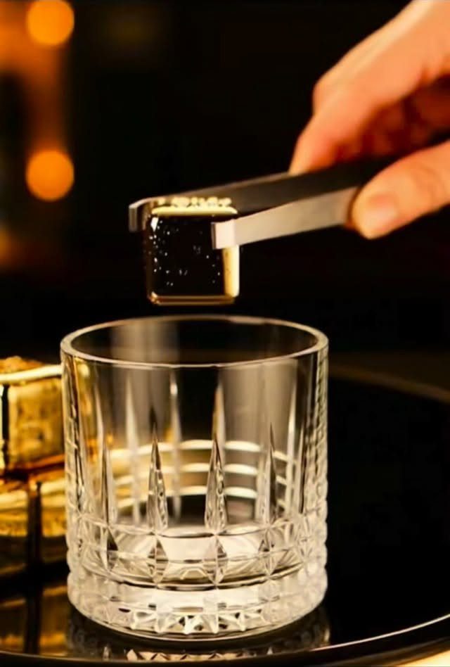 Gold whiskey stones being placed into a crystal glass with metal tongs