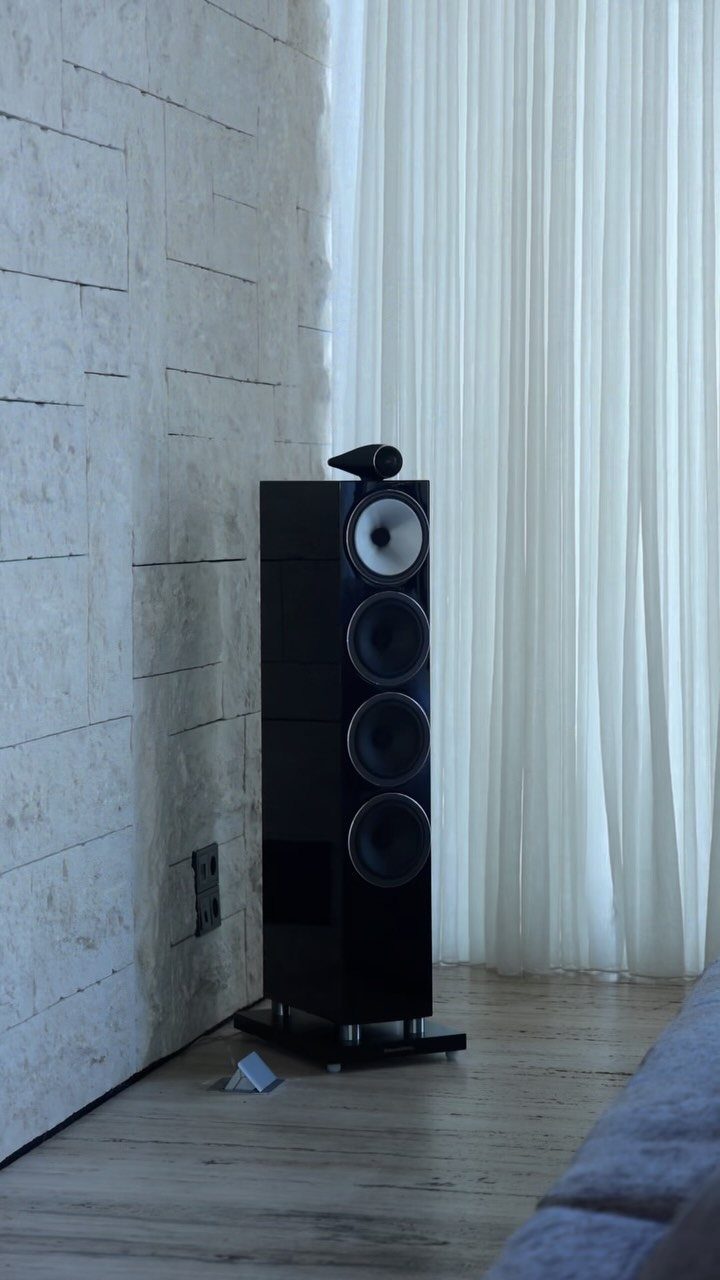 Tall black floor-standing speaker against a stone wall and white curtains.