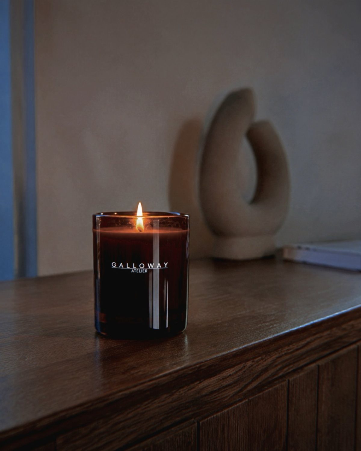 Lit scented candle in a dark glass jar on a wooden dresser.