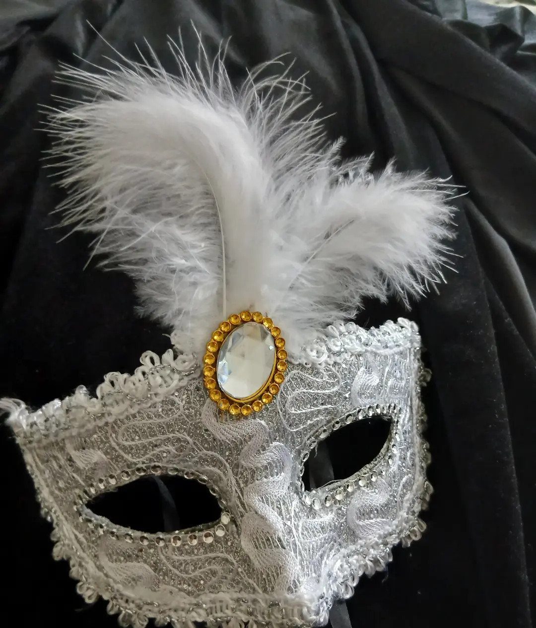 White lace masquerade mask with gold jewel and white feathers on black fabric