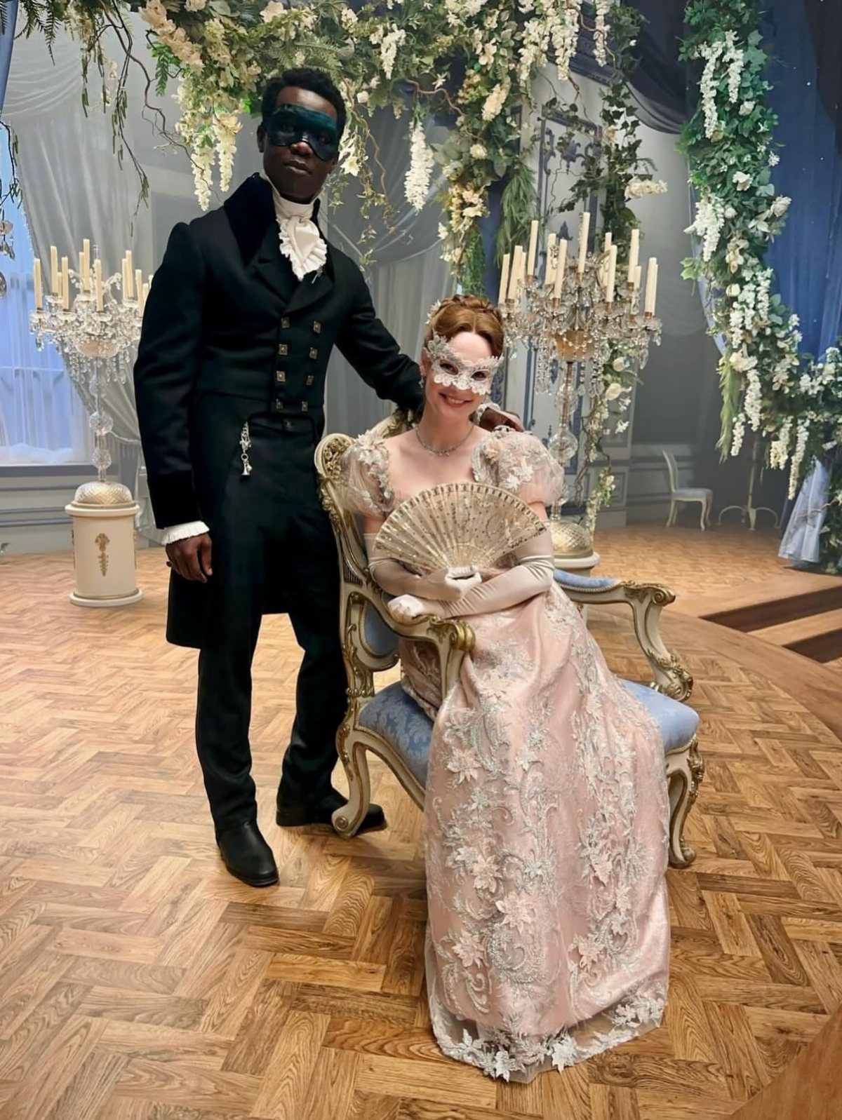 Woman in pink lace ball gown and man in black suit wearing masquerade masks.