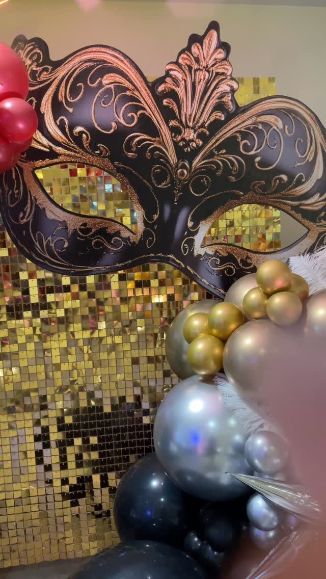 Ornate black and gold masquerade mask cutout with gold sequin backdrop and balloons.