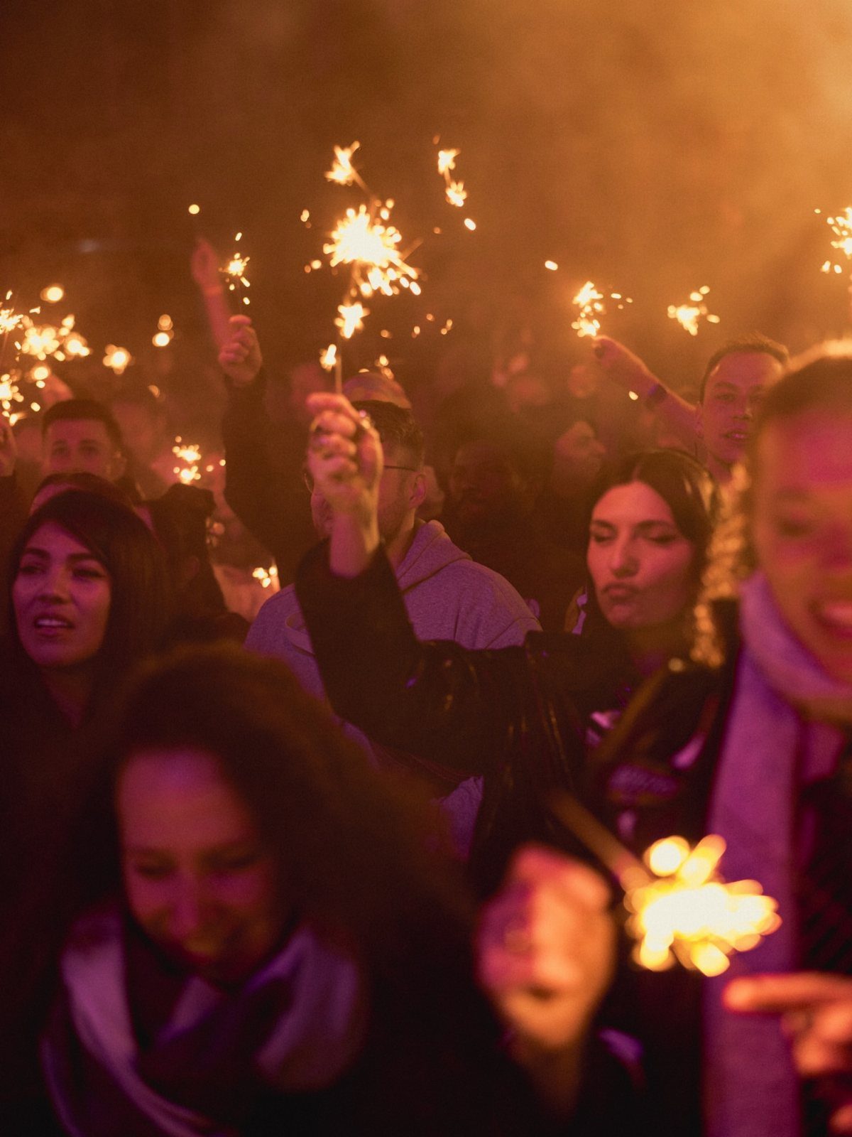 Crowd of people holding lit sparklers during a nighttime celebration with orange lighting.