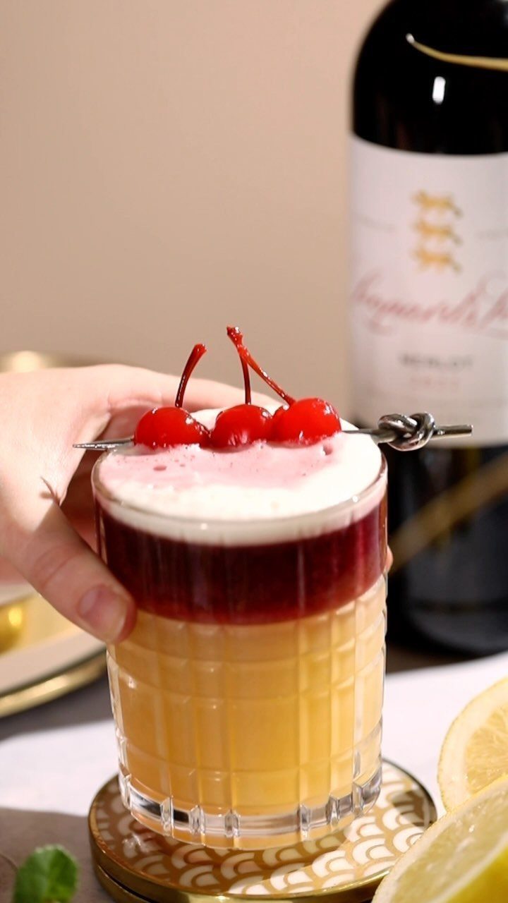 Layered cocktail with cherries and foam held in front of a wine bottle.