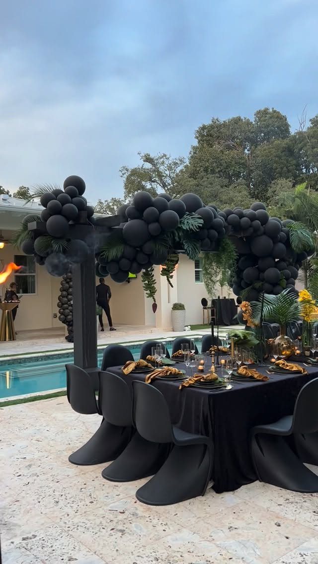 Black balloon garland arch framing a black dinner table by a swimming pool.