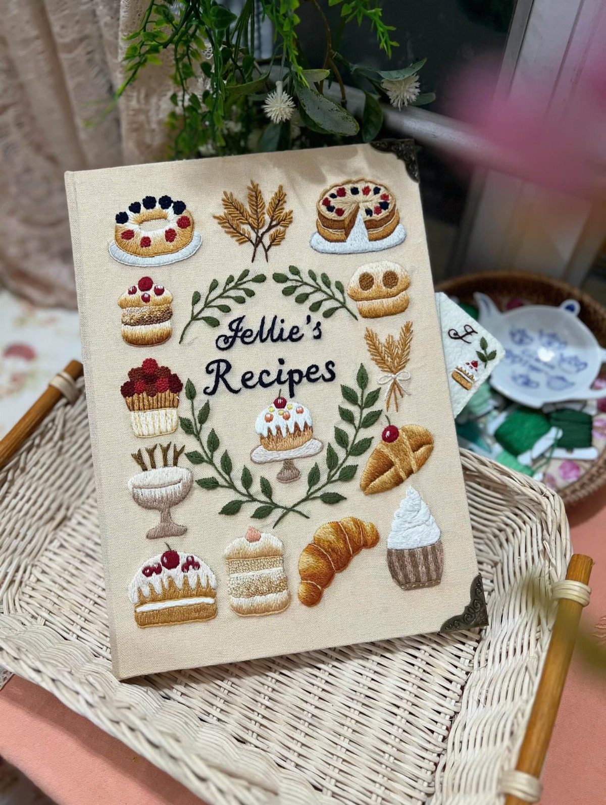 Embroidered recipe book with cakes and pastries on a white wicker tray.