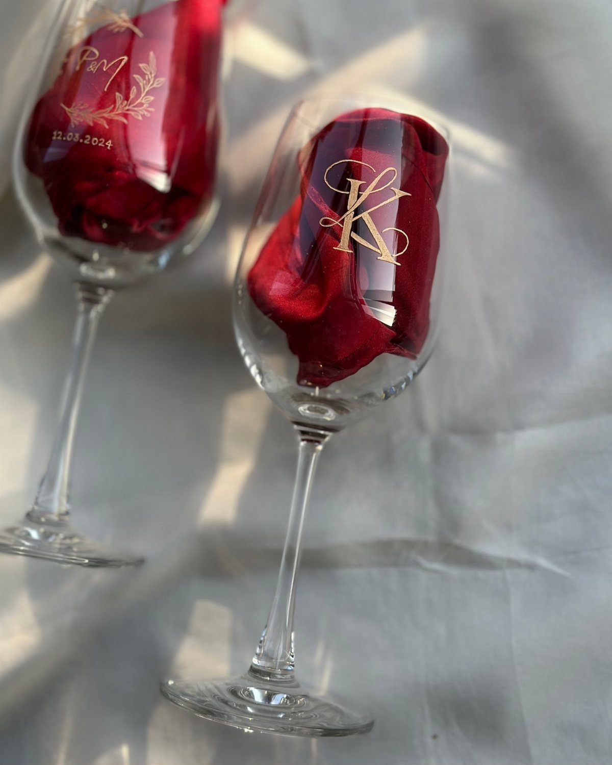 Two wine glasses with gold engraved initials containing red rose petals on white fabric