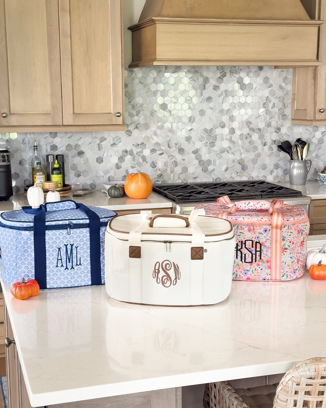 Three monogrammed insulated cooler bags on a white kitchen counter with small pumpkins.