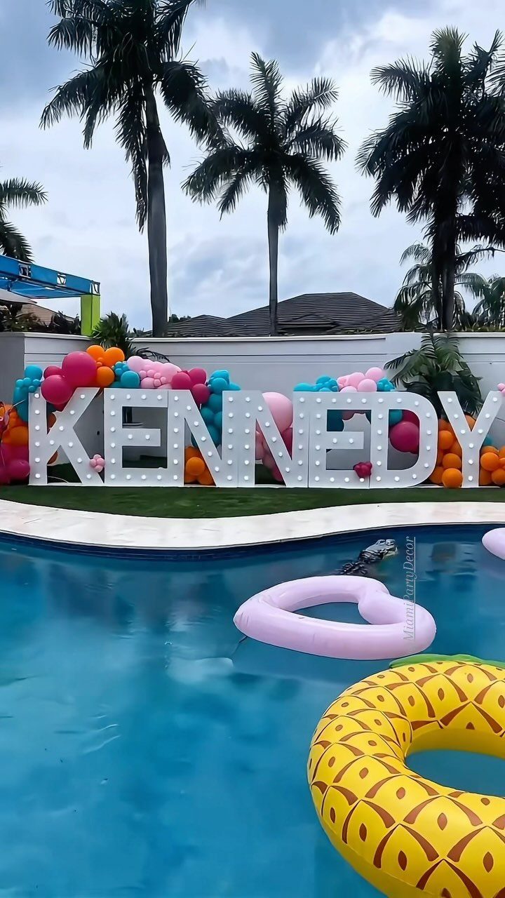 Pool party decorations featuring large illuminated letters, colorful balloons, and floating pool rings.