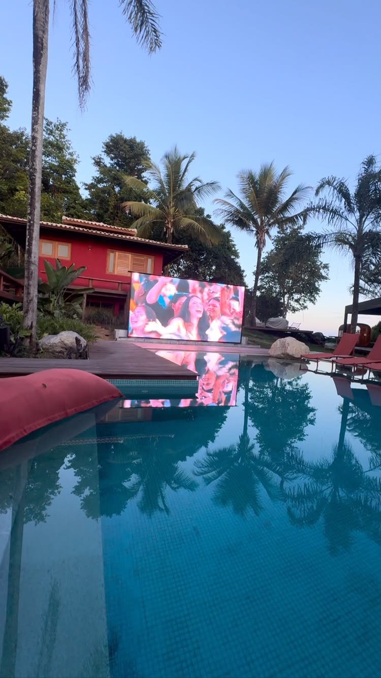 Outdoor movie screen by a swimming pool with palm trees and red house.