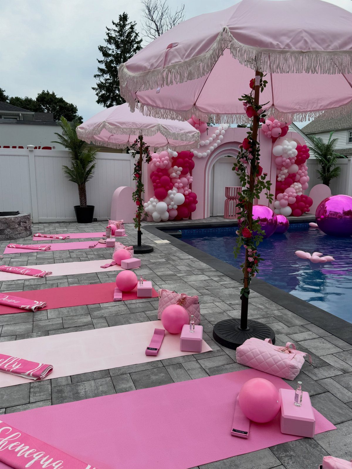 Pink poolside party decorations with yoga mats, umbrellas, and balloon arches by a pool.