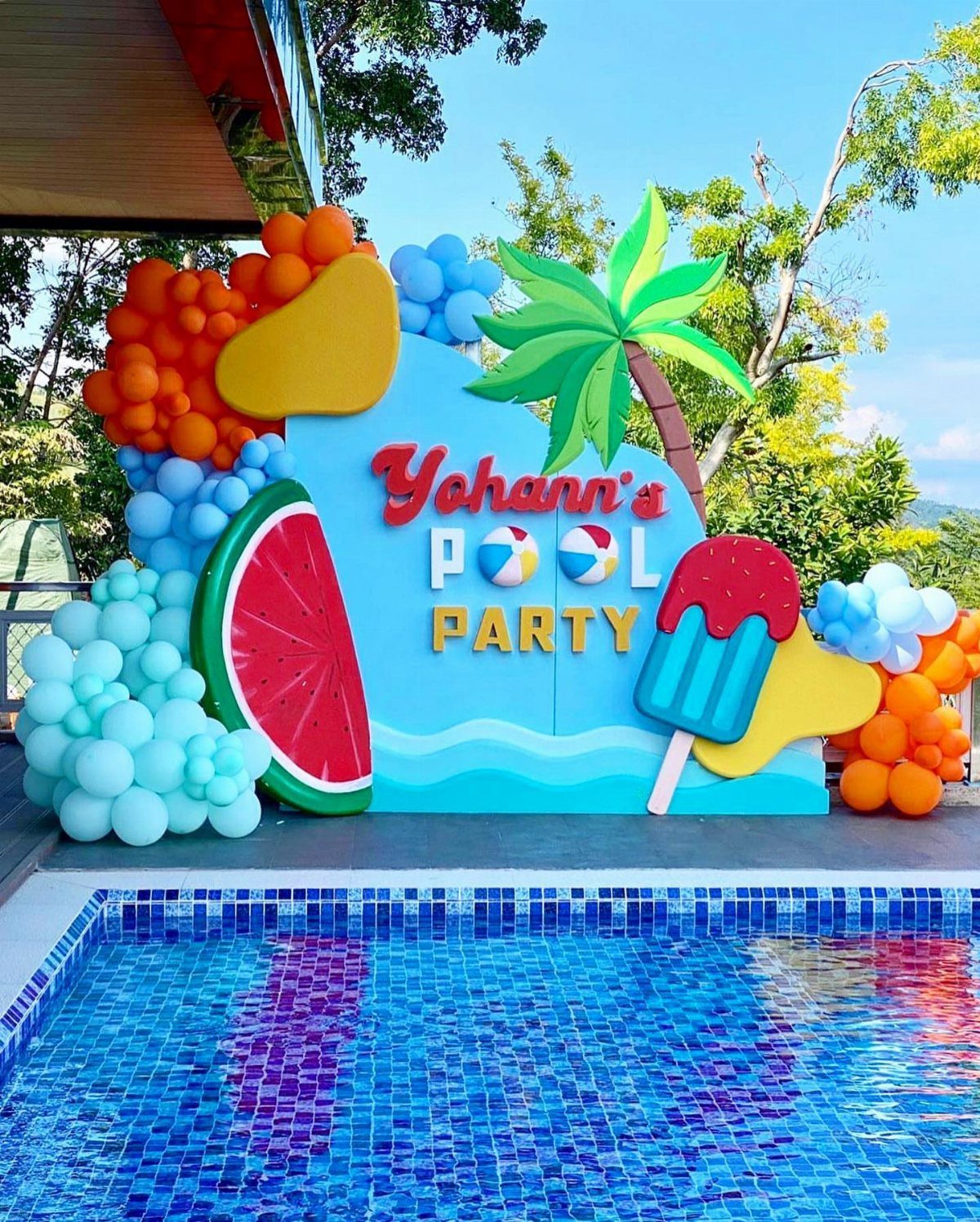 Colorful pool party welcome banner with balloons, fruit cutouts, and a palm tree.