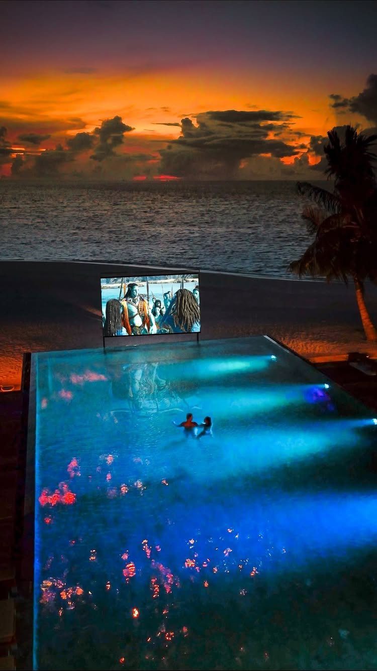 Outdoor movie screen by a glowing blue pool during a tropical sunset.