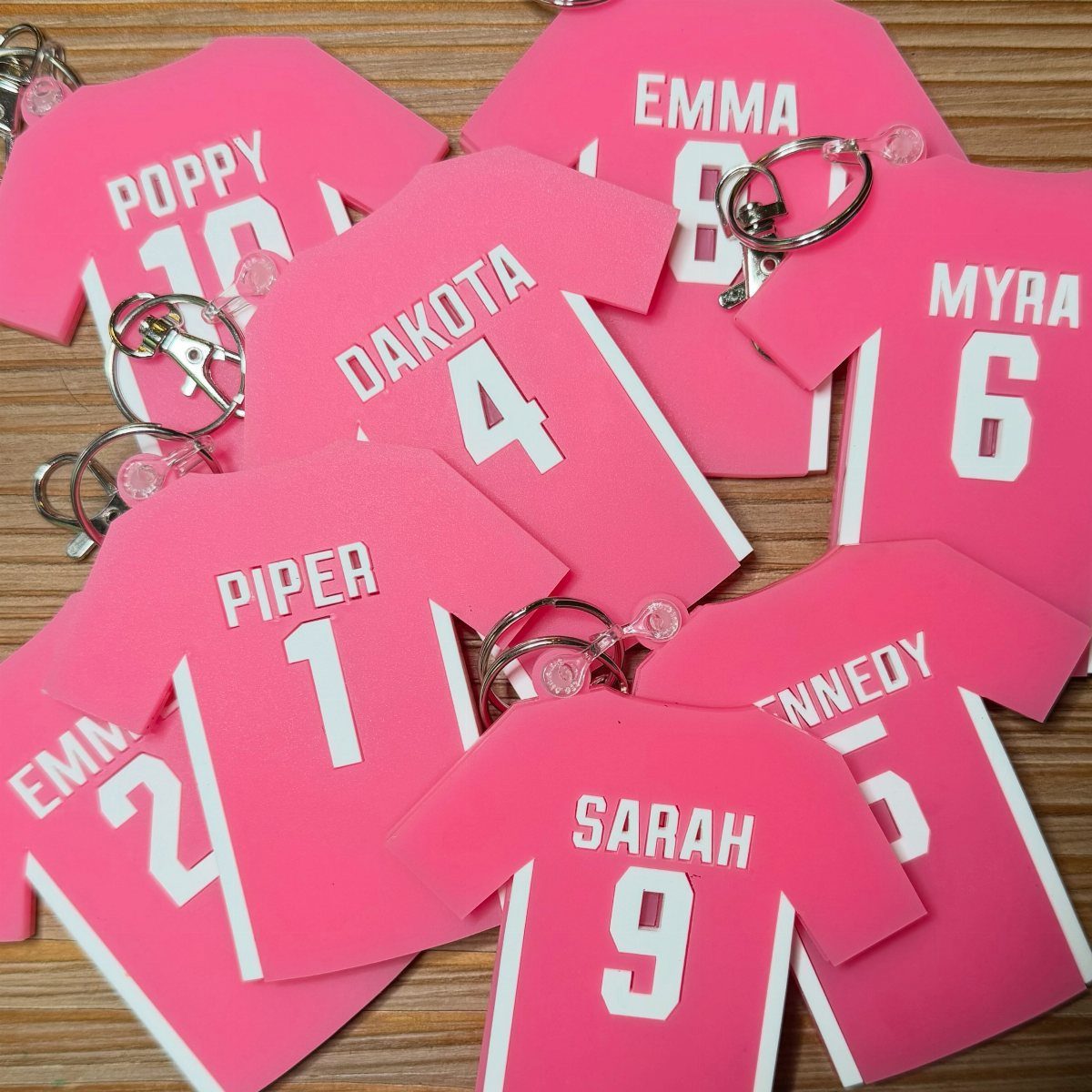 Pink jersey-shaped personalized bag tags with names and numbers on a wooden surface.