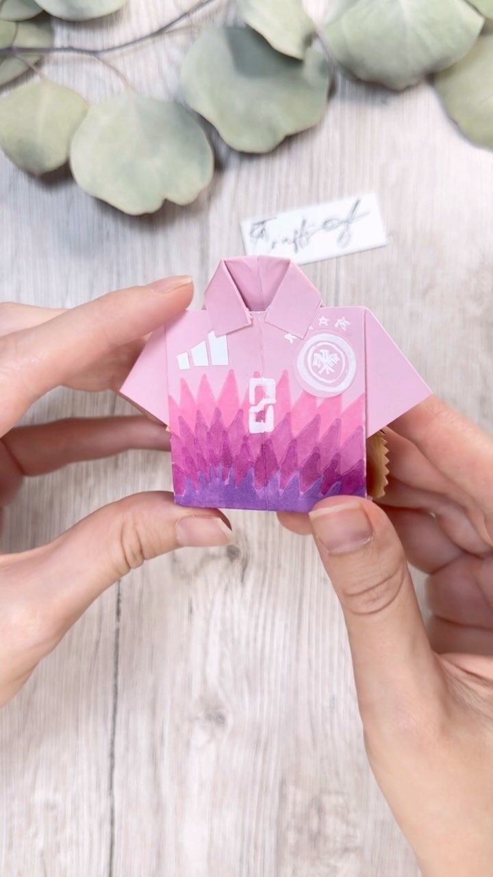 Small pink paper favor box shaped like a soccer jersey held by hands.