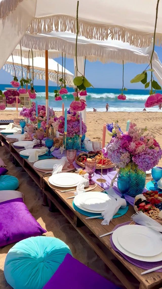 Beach party table setting with purple flowers, blue glassware, and colorful floor cushions.