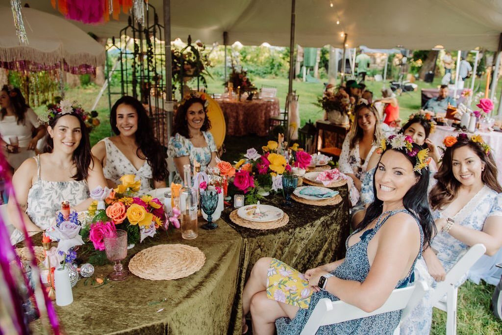 Women with flower crowns sitting at a decorated table under a white party tent.