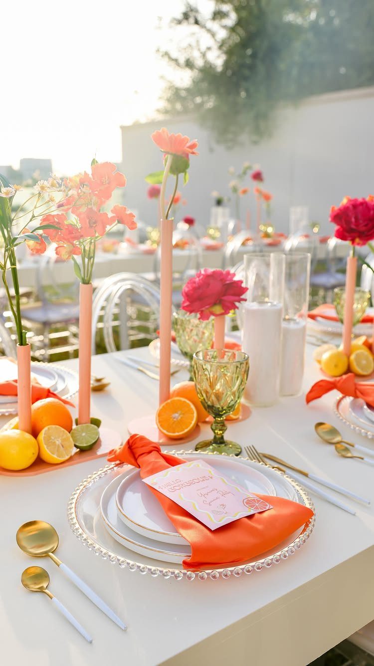 Outdoor table setting with orange napkins, citrus fruits, and peach-colored floral arrangements.