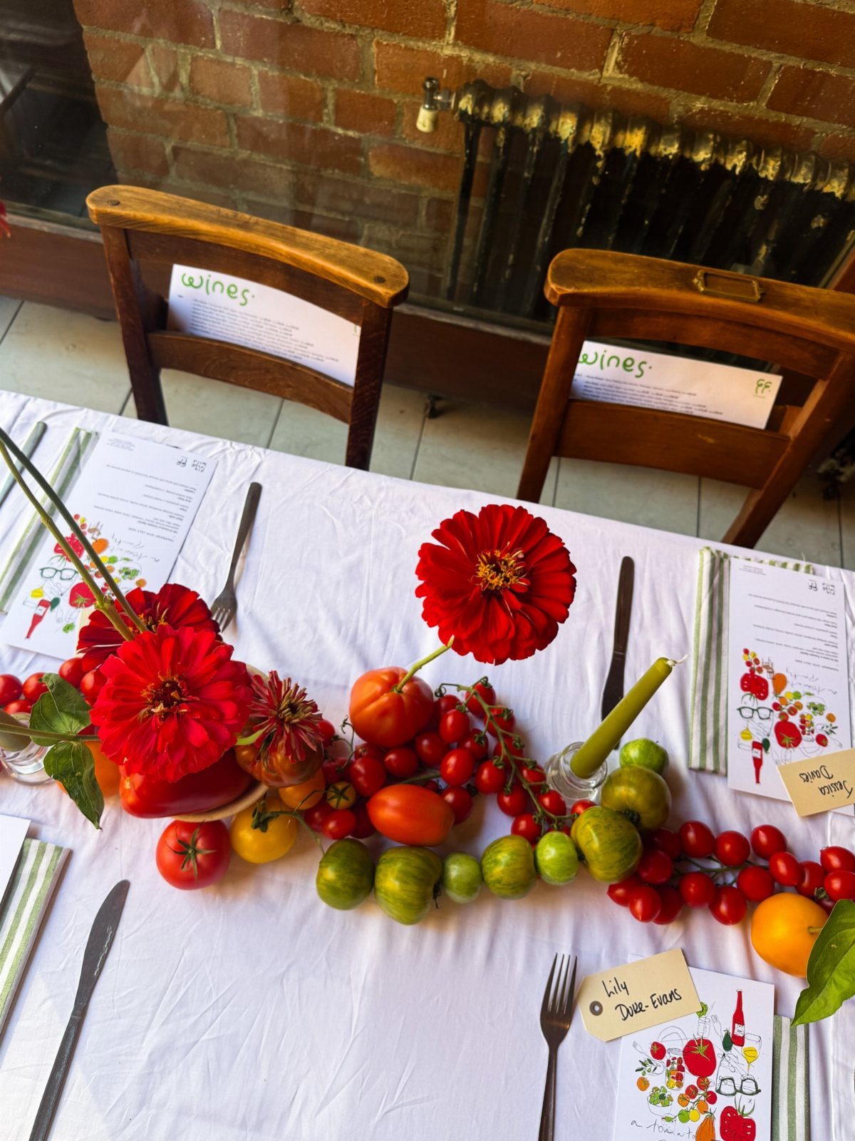 Table setting with a centerpiece of fresh tomatoes and red flowers on white cloth.