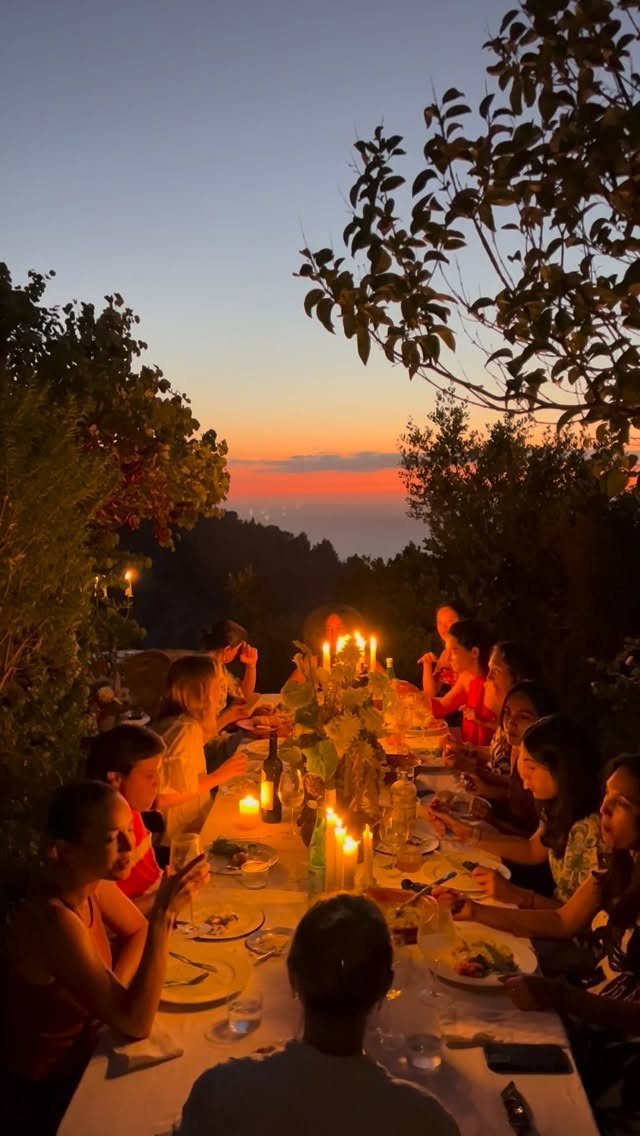 People dining at a long candlelit table outdoors during a vibrant orange sunset.
