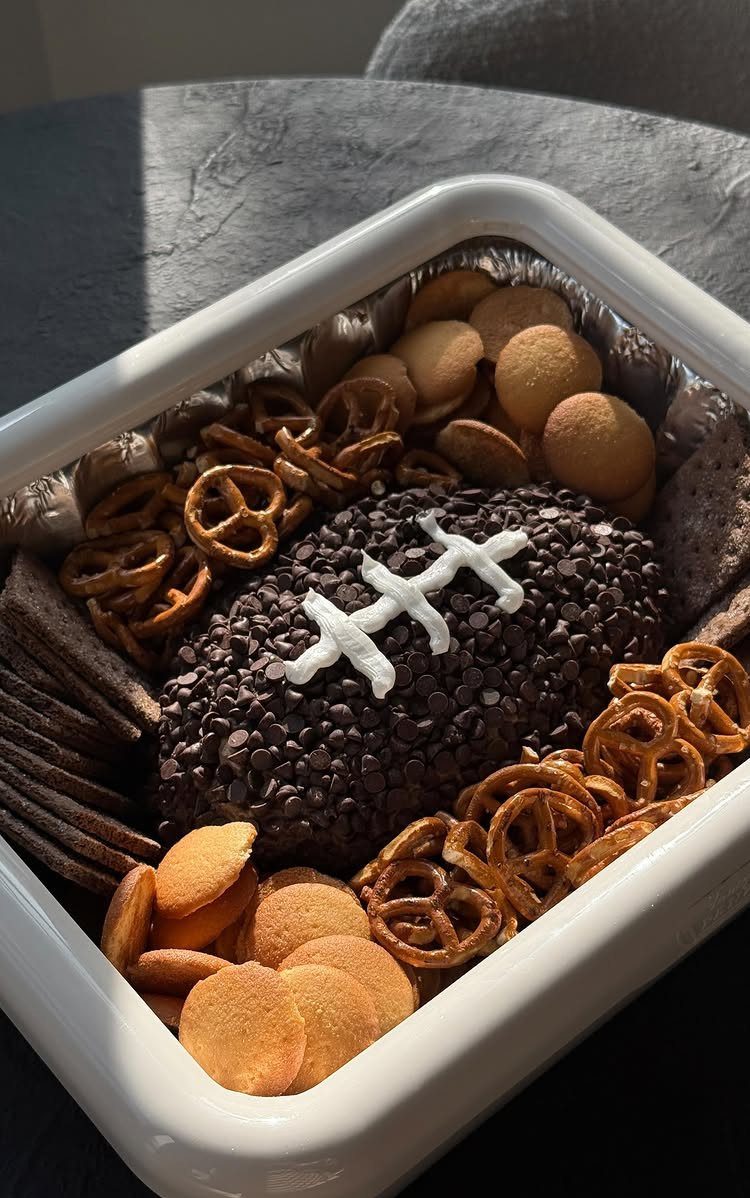 Football-shaped chocolate dip surrounded by pretzels and crackers in a white tray.