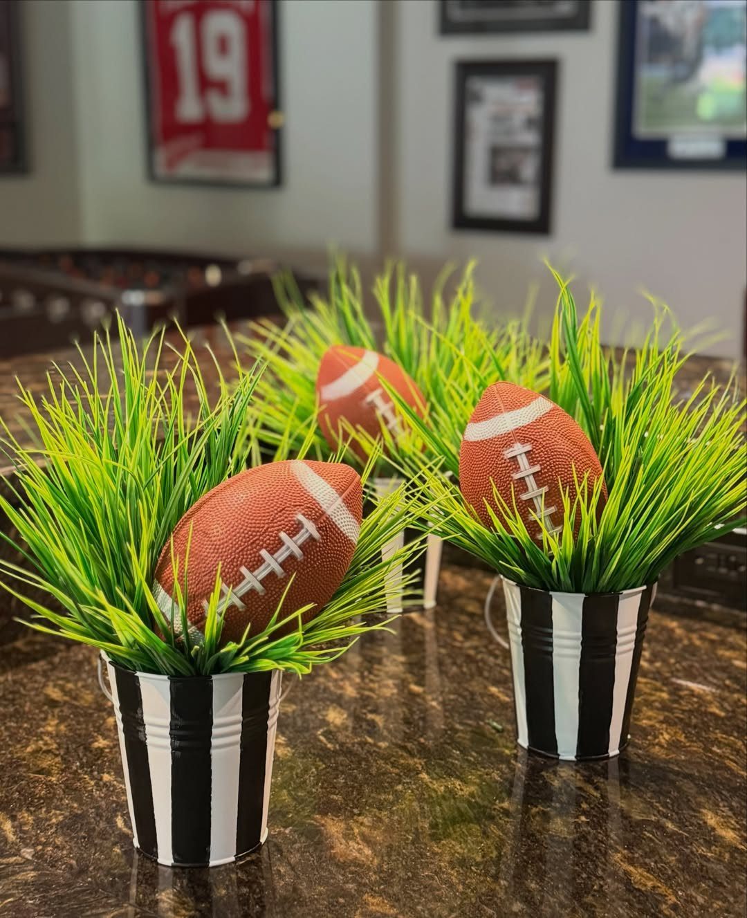 Three footballs nestled in green grass inside striped buckets on a granite counter.
