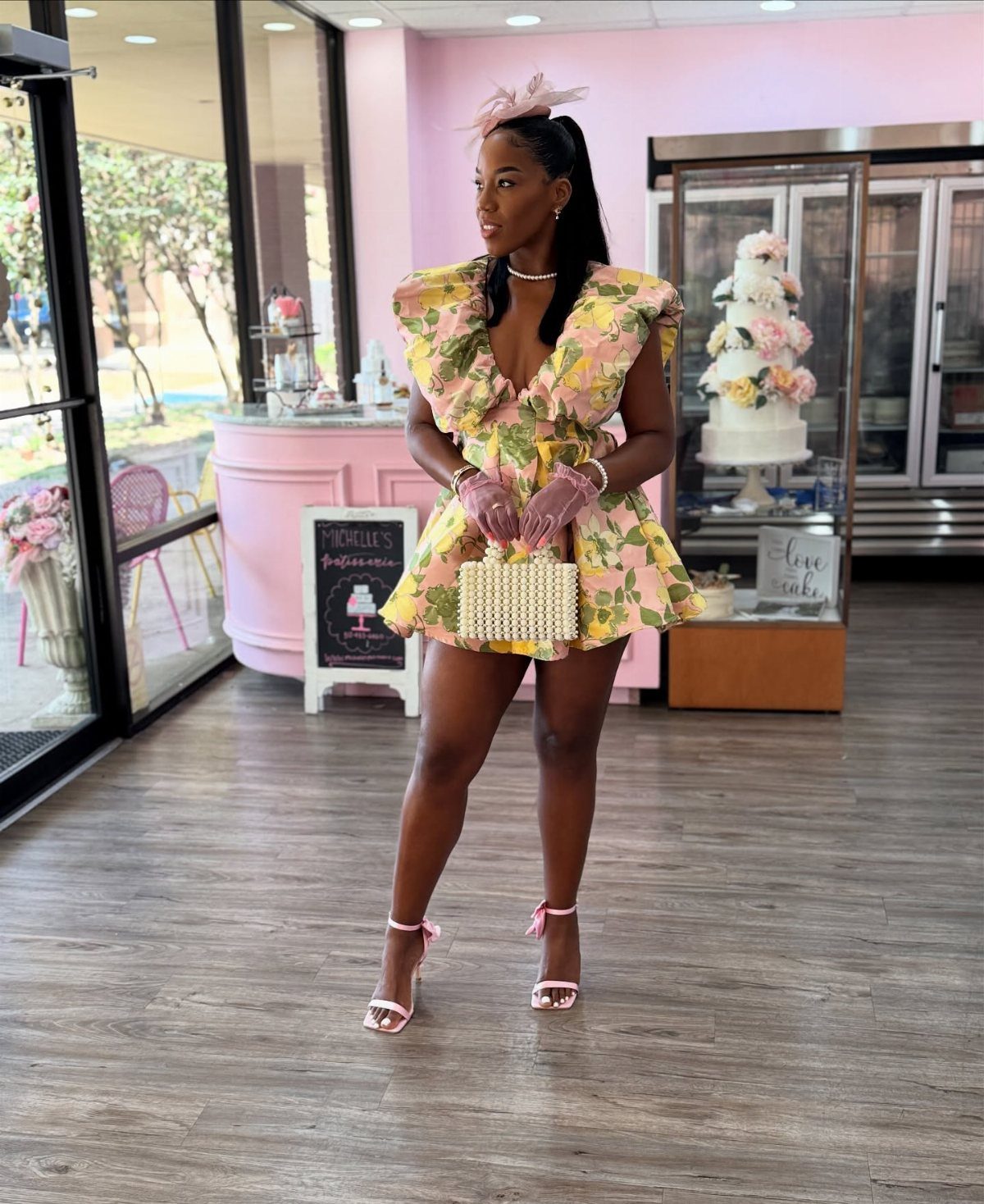 Woman wearing a yellow floral dress, pink gloves, and heels in a bakery.