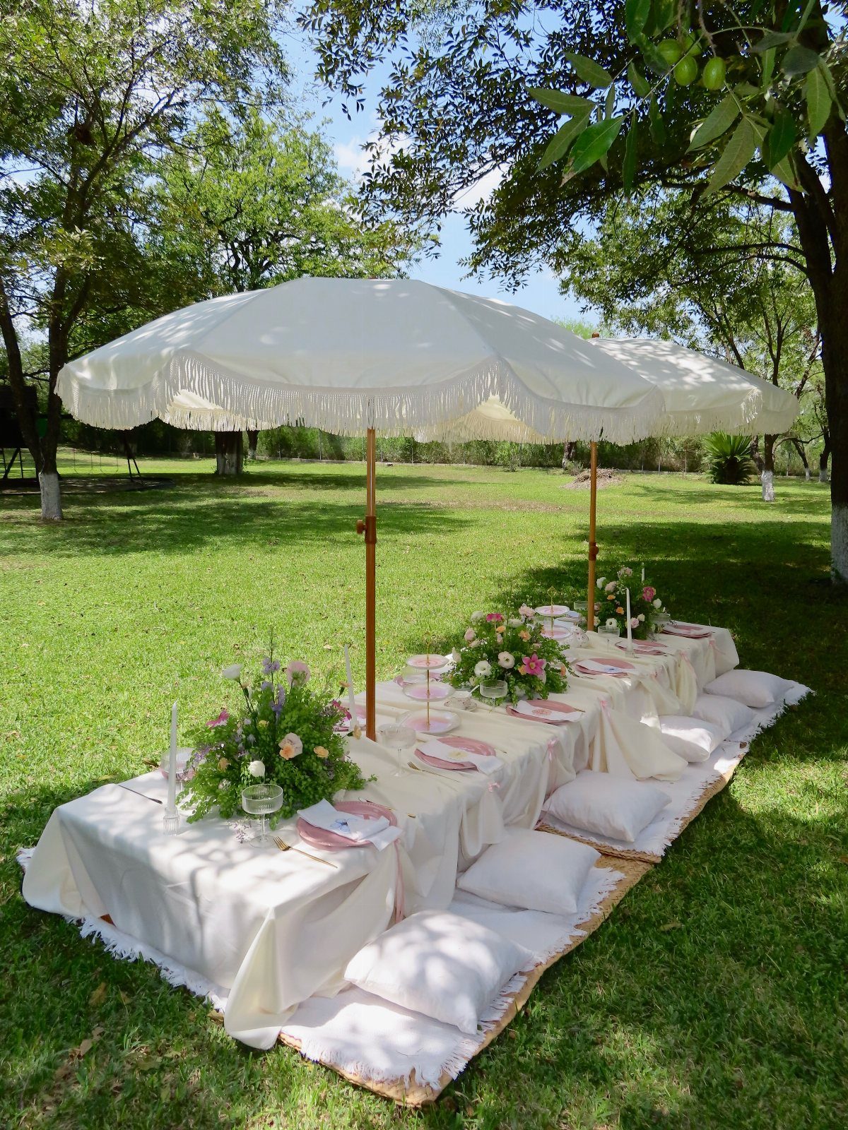 Low white table with floral centerpieces and cushions under umbrellas in a garden