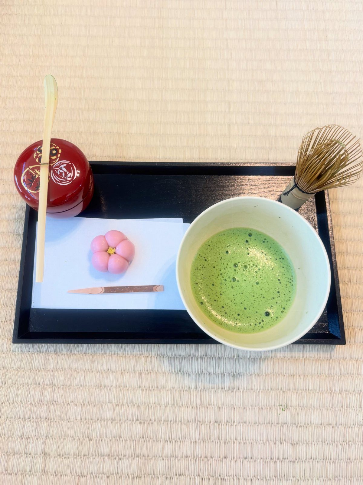 Matcha tea in a white bowl with a pink sweet and bamboo whisk.