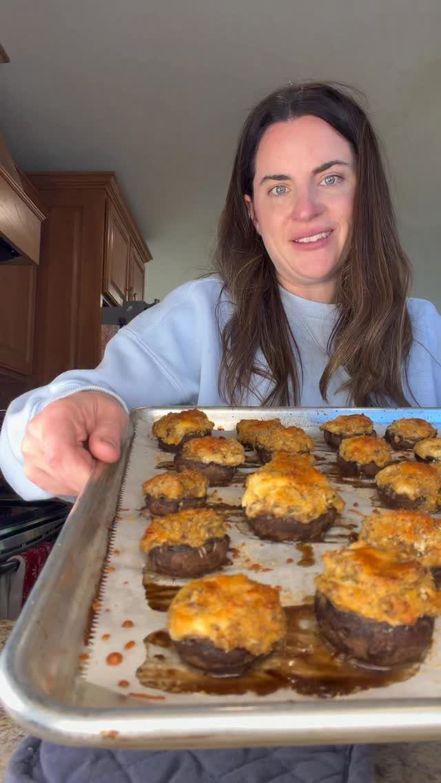 Woman holding a baking sheet of golden-brown cheesy stuffed mushrooms in a kitchen.