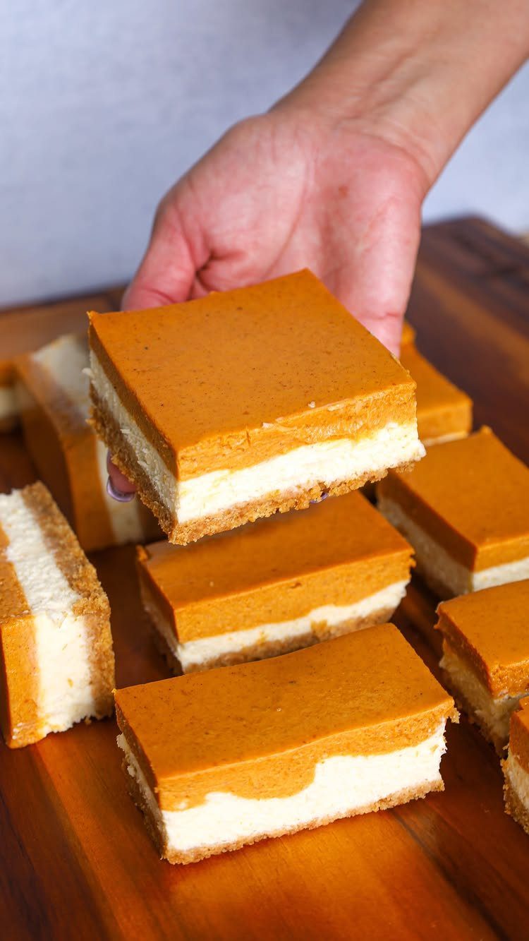 Layered pumpkin cheesecake squares on a wooden board, one held by a hand.