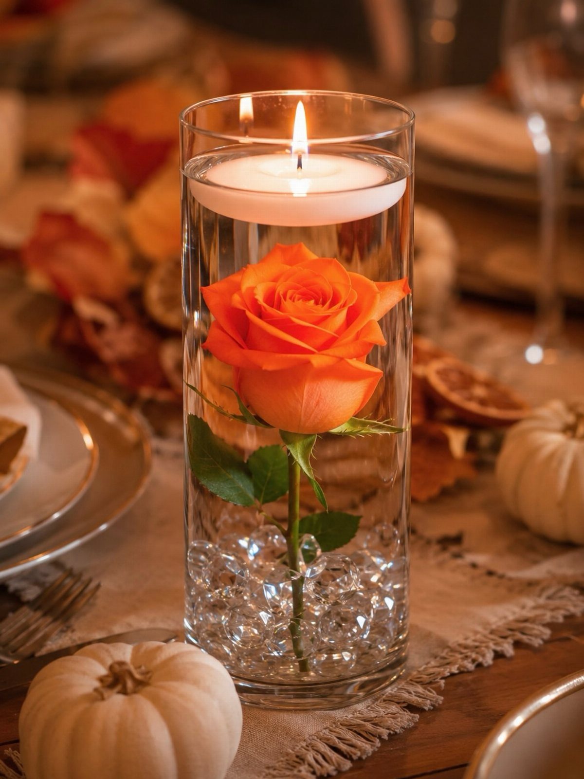 Floating white candle atop an orange rose in a glass cylinder vase centerpiece.