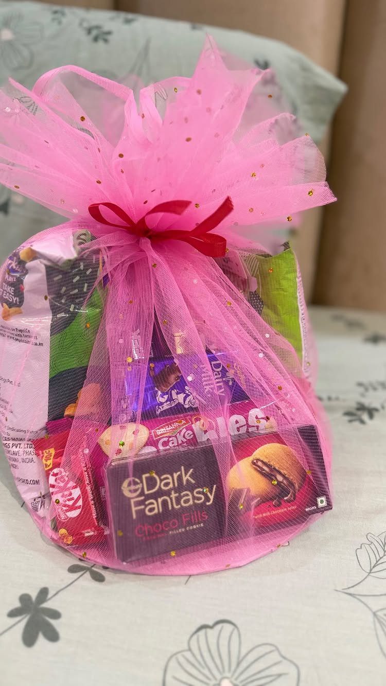 Gift basket wrapped in pink tulle with red ribbon containing assorted chocolate snacks.