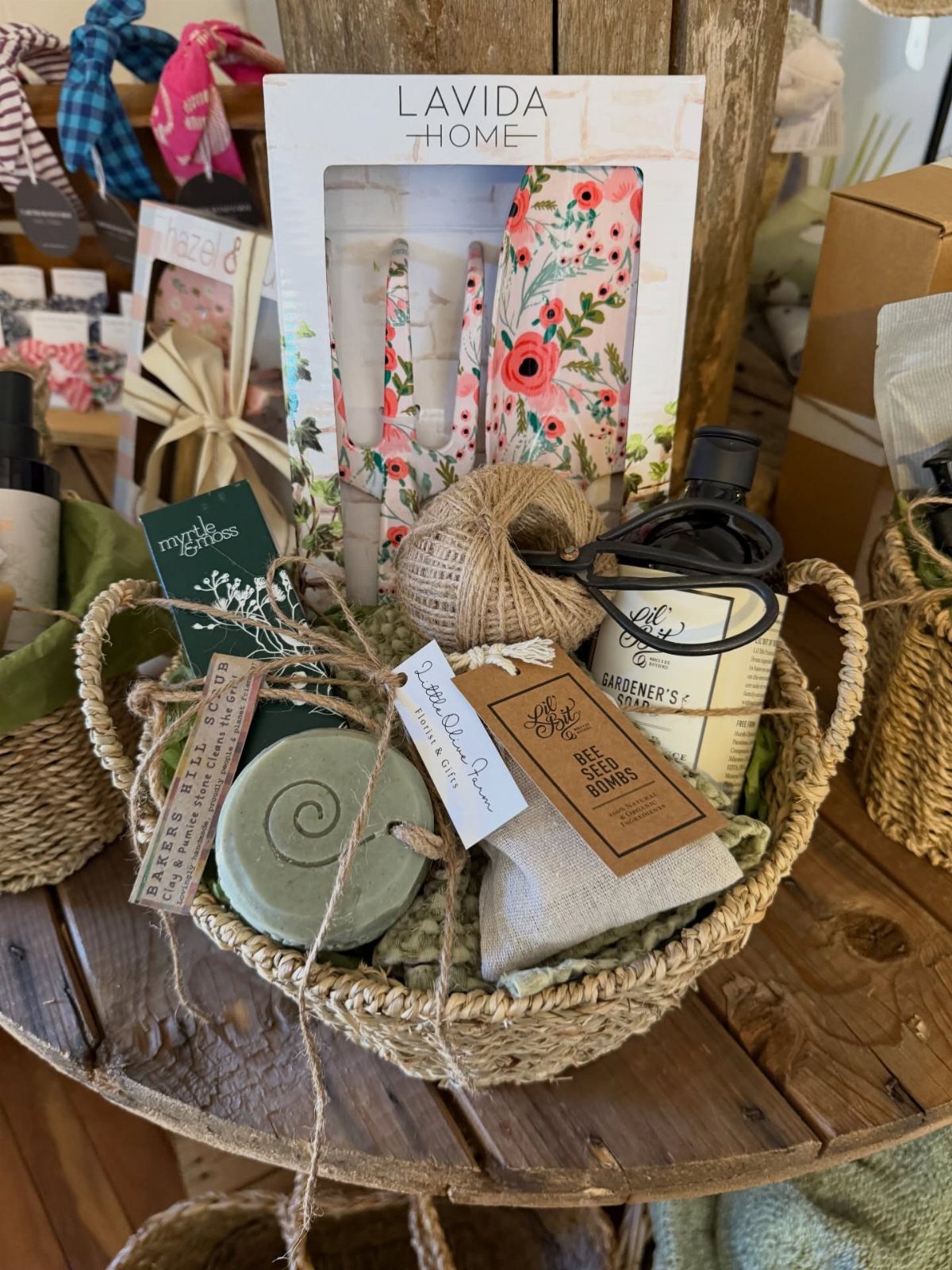 Woven gift basket containing gardener's soap, floral napkins, and seed bombs on wood.