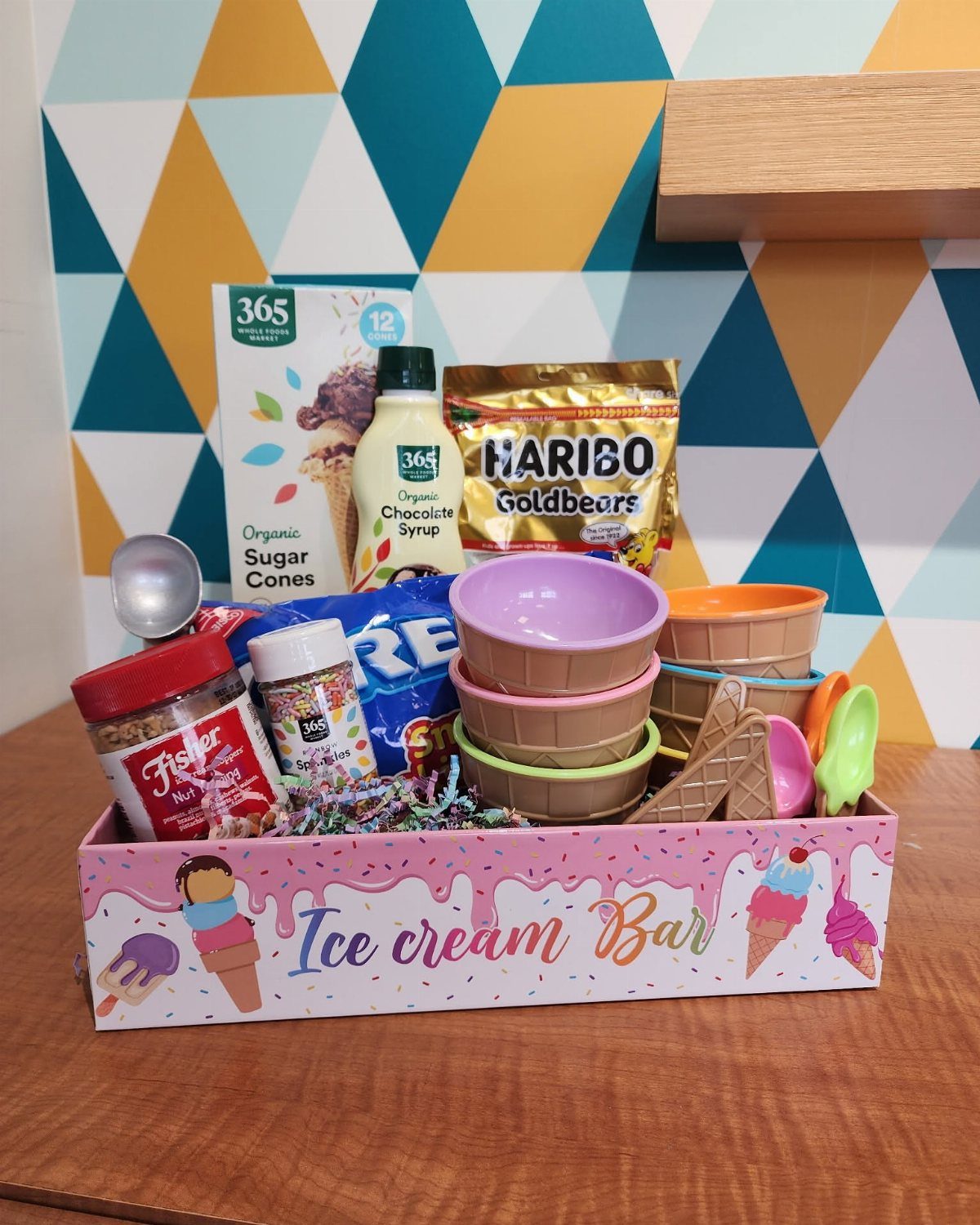 Ice cream gift basket with cones, bowls, and toppings in a pink tray.