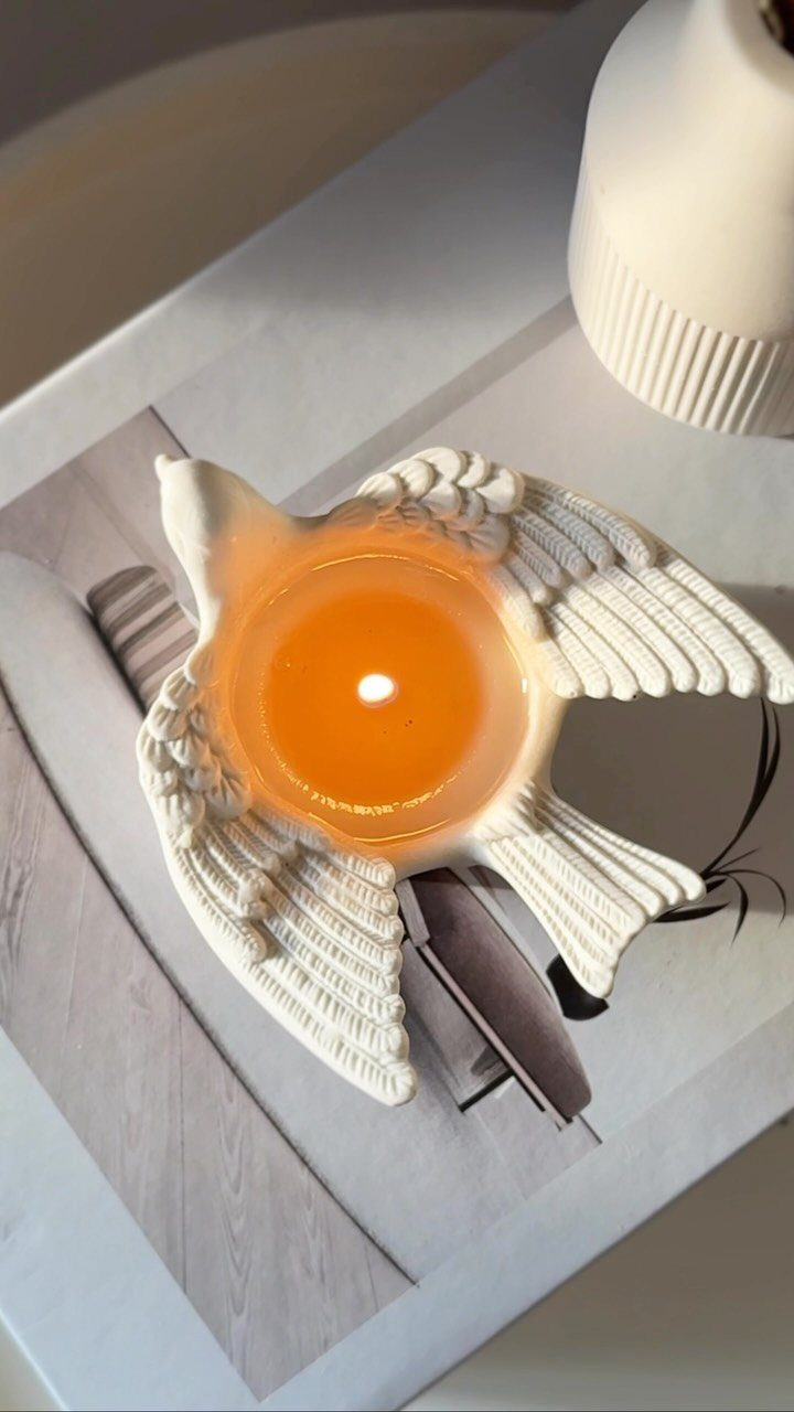 Lit orange candle in a white ceramic bird-shaped holder on a light surface.
