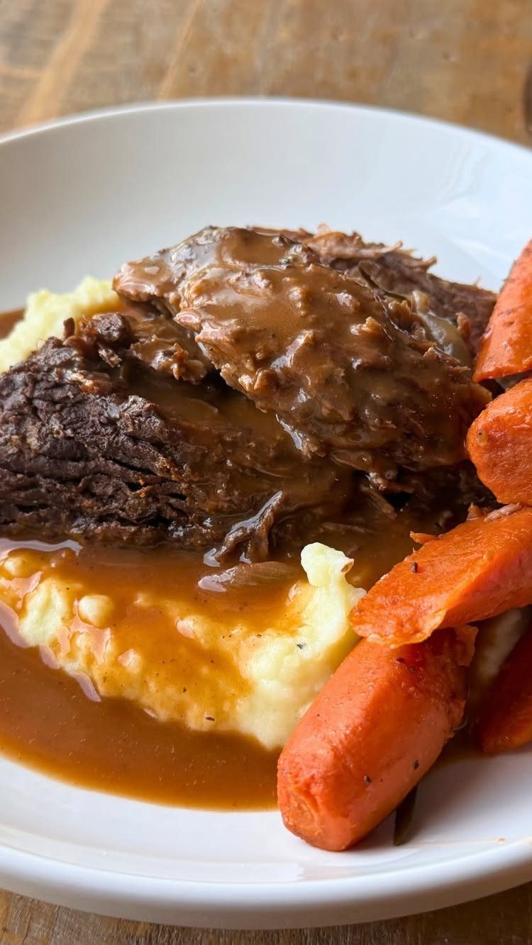 Pot roast with brown gravy, mashed potatoes, and glazed carrots in a white bowl.