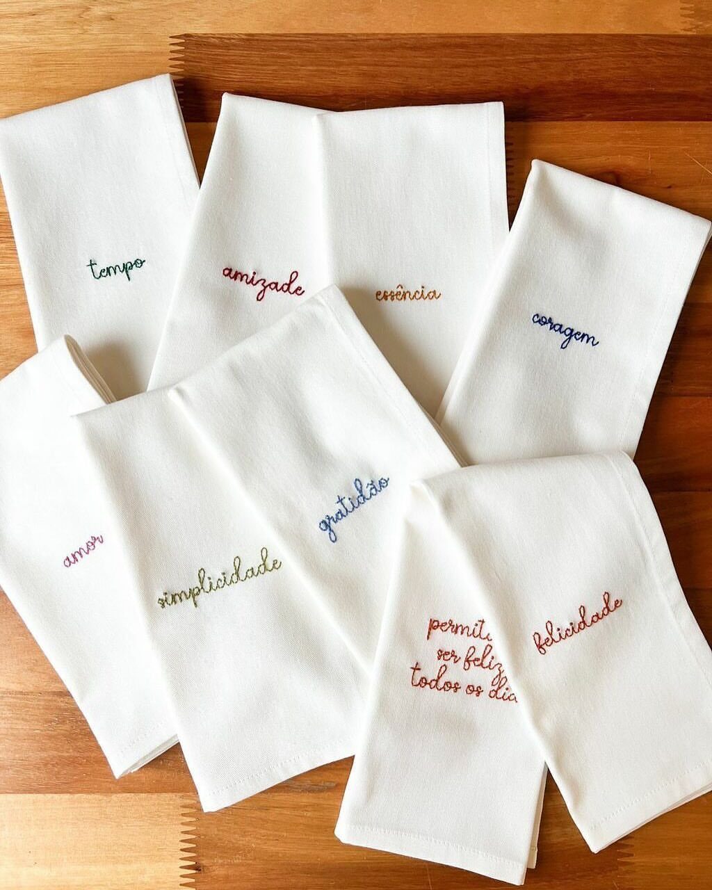 White kitchen towels with colorful embroidered Portuguese words on a wooden table.