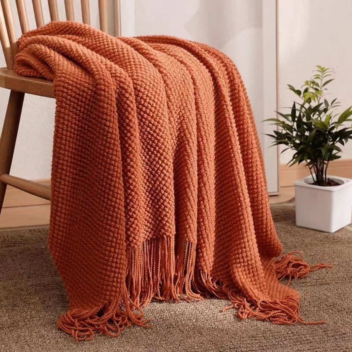 Rust-orange textured throw blanket draped over a wooden chair next to a potted plant.