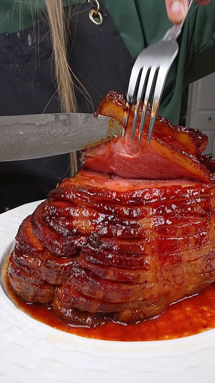 Glazed spiral-sliced ham on a white platter being carved with a knife and fork.