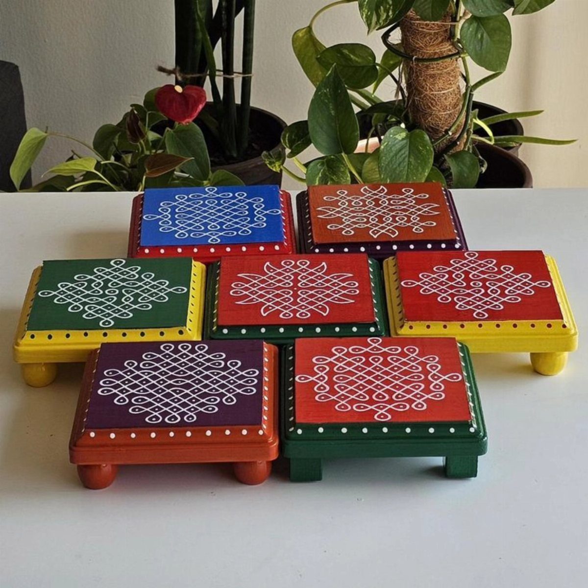 Colorful wooden coasters with white patterns arranged on a white table near plants.