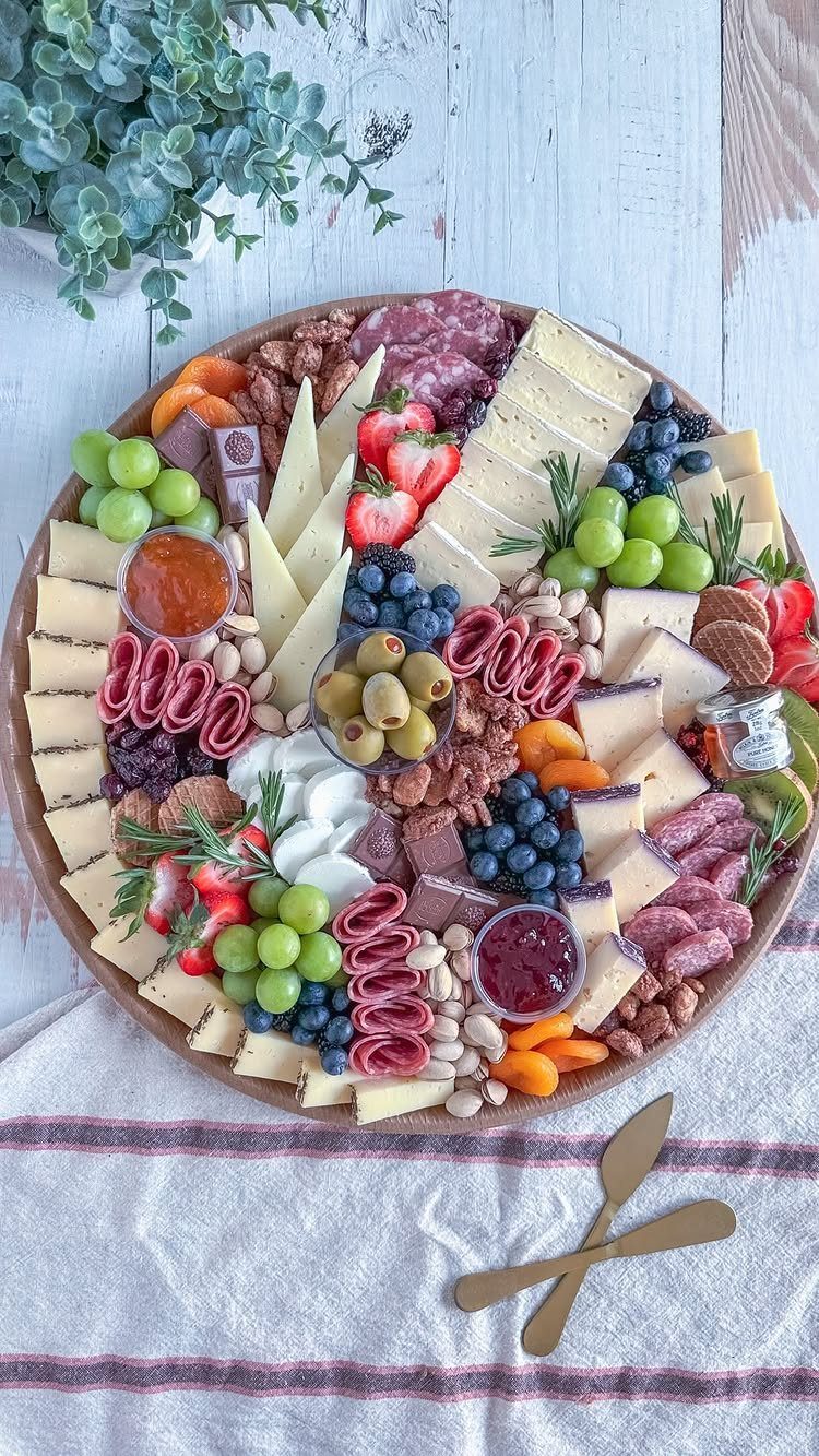 Round wooden charcuterie board loaded with cheeses, meats, fruits, and nuts on white wood.