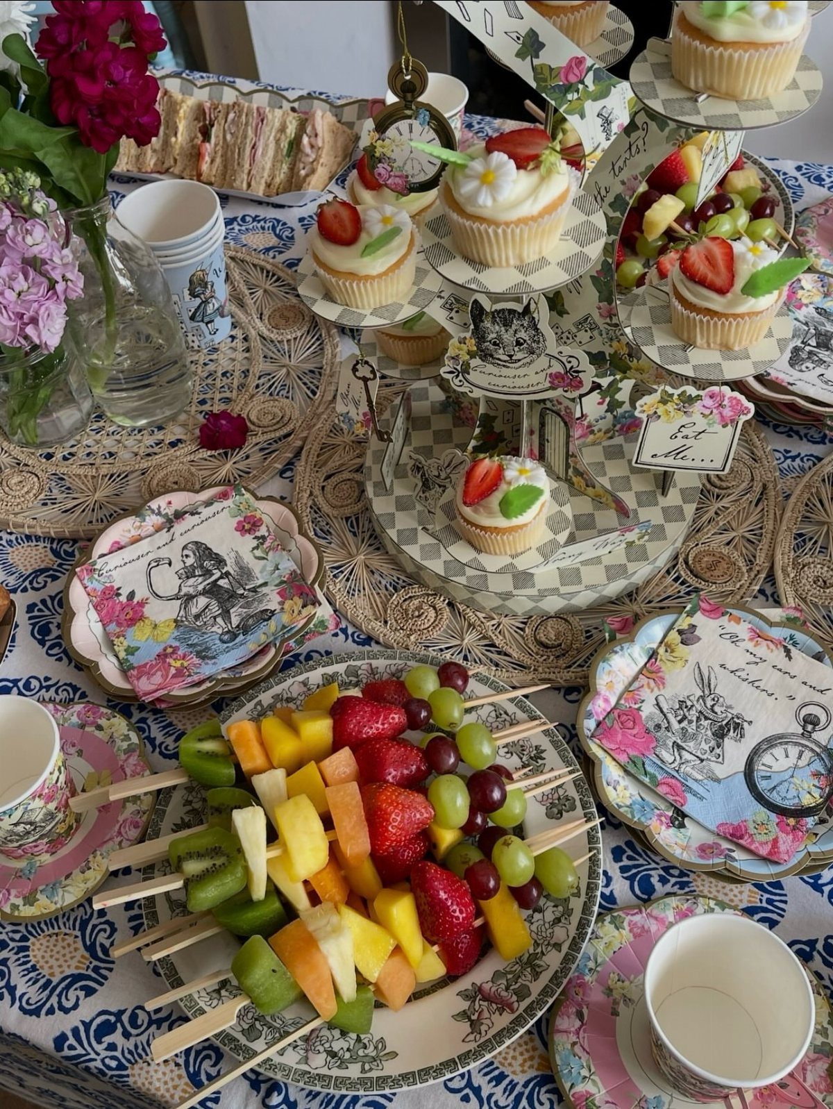 Alice in Wonderland themed tea party table with cupcakes, fruit skewers, and floral napkins.