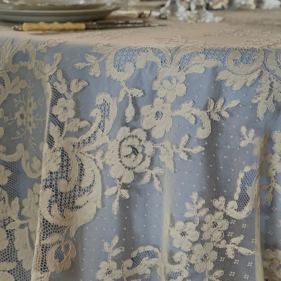 Cream floral lace tablecloth overlaying a light blue fabric on a set table.