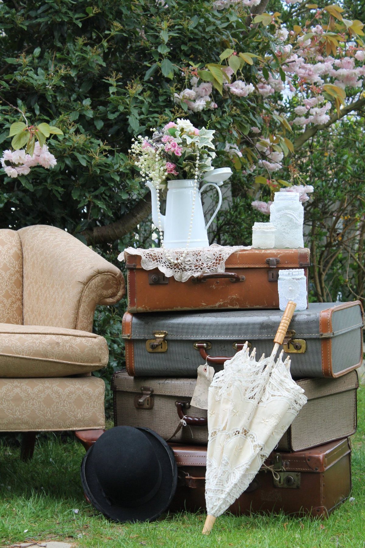 Stacked vintage suitcases topped with flowers and a white pitcher in a garden setting.