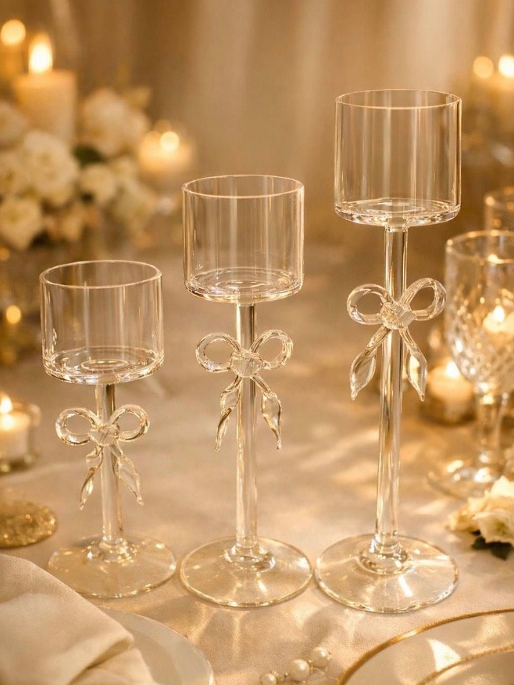 Three clear glass candle holders with bows on a white wedding table.