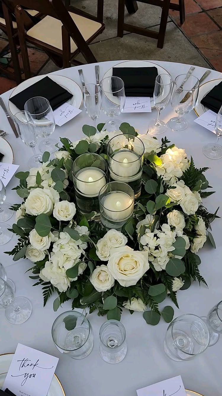 Floating white candles in glass cylinders surrounded by white roses and green foliage.